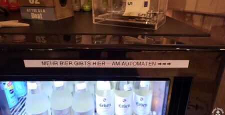 Ein Kühlschrank voller Glasflaschen Erdelt-Bier. Darüber hängt ein Schild mit der Aufschrift „Mehr Bier gibt’s hier – am Automaten“ und nach rechts zeigenden Pfeilen, was darauf hinweist, dass in einem nahegelegenen Automaten noch mehr von diesem ehrlichen Getränk erhältlich ist – perfekt für eine spätere Stunde.