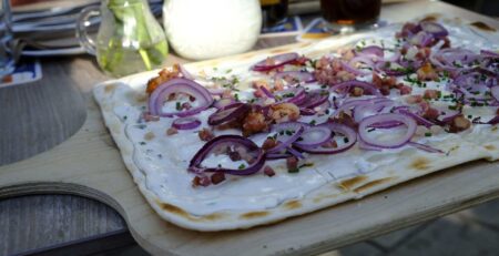 Ein frisch gebackener Flammkuchen mit cremiger weißer Soße, knusprigen Speckstücken, dünn geschnittenen roten Zwiebeln und gehackten grünen Kräutern wird auf einem Holzbrett serviert. Dieses leckere Gericht wird auf einem Tisch präsentiert, im Hintergrund stehen eine Vase mit Blumen, Getränken und Besteck.