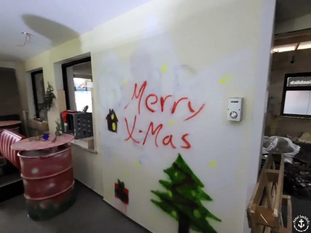 Auf einer Innenwand ist in roten Buchstaben ein Graffiti mit der Aufschrift „Merry X-Mas“ zu sehen. Darunter ist ein kleines Haus mit Rauch, einem geschmückten Weihnachtsbaum und einem kleinen Geschenk gezeichnet. Im Vordergrund ergänzen ein rotes Fass und unfertige Baumaterialien die wuselig anmutende Szenerie, die an einen geschäftigen Familienbetrieb erinnert.