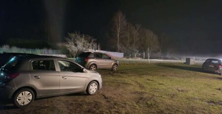 Drei Autos sind nachts auf einer Wiese geparkt, ihre Positionen lassen auf eine informelle Anordnung schließen. Der schwach beleuchtete Hintergrund zeigt Bäume und einen Zaun. Die Szene ist ruhig und still, erinnert an ein Gemälde aus Der Sturm, ohne sichtbare Menschen oder Aktivitäten, die die Stille stören.