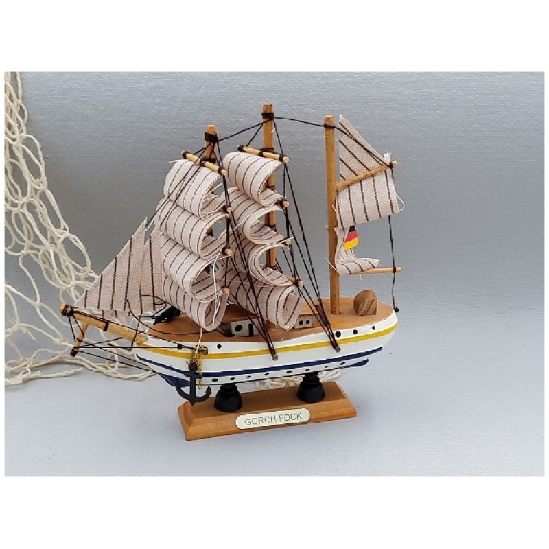 Ein detailliertes Miniaturmodell der Gorch Fock aus ca. 16 cm Holz steht auf einer grauen Oberfläche. Das Modell verfügt über drei Masten mit gestreiften Segeln, einen gelb und blau linierten Rumpf und eine kleine deutsche Flagge. Im Hintergrund ist teilweise ein Fischernetz zu sehen.