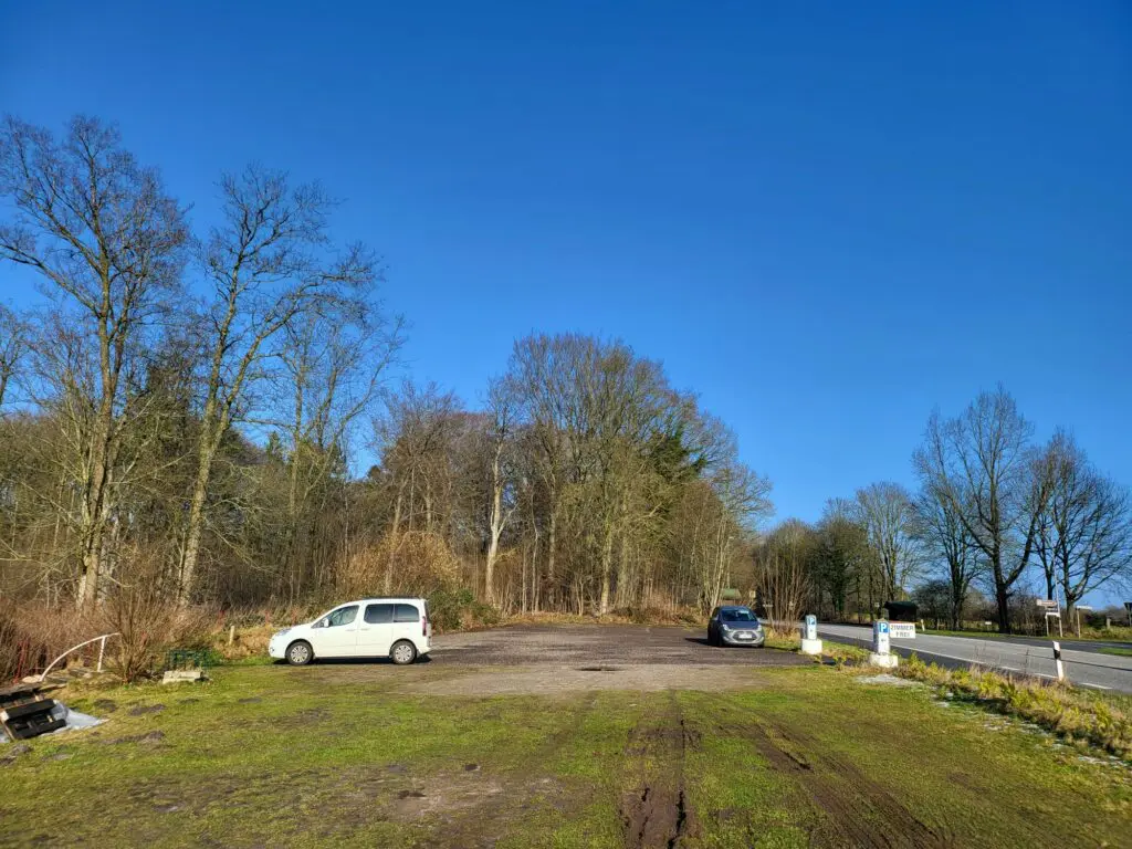 Ein klarer, sonniger Tag auf einem Parkplatz mit zwei Autos auf einem Feld mit Erde und Gras. Um den Parkplatz herum stehen blattlose Bäume und trockene Büsche. Nach rechts verläuft eine asphaltierte Straße, daneben ein kleines Gebäude und Poller. Der Himmel ist strahlend blau und bildet einen Kontrast zum matschigen Rasen darunter.