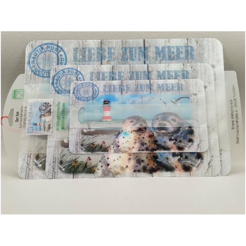 Eine Präsentation mehrerer Pakete mit einem Leuchtturm und zwei Robben an einem Sandstrand. Die Verpackung trägt den Text „Schneidbrett 3er-Set Liebe zum Meer“ und enthält ein Röhrchen und Muscheln. Der Hintergrund imitiert verwitterte Holzbretter und erinnert an ein altes Schneidbrett.