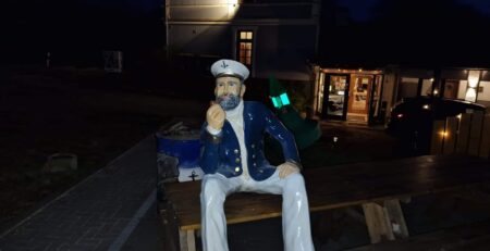 Eine Statue eines sitzenden Matrosen mit der Aufschrift „Ahoi Hein!“ auf seinem Hut, einer blauen Uniform und einem weißen Bart sitzt auf einem hölzernen Picknicktisch. Der Matrose scheint eine Pfeife zu halten. Im Hintergrund ist ein Gebäude zu sehen, das nachts mit verschiedenen Lichtern beleuchtet wird.