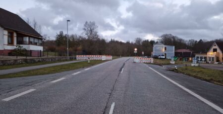 Eine Straße ist mit Verkehrsbarrieren und Warnschildern gesperrt. Auf beiden Seiten der Straße stehen Häuser. Der Himmel ist bewölkt und bedeckt, im Hintergrund sind Bäume zu sehen. Die Szene, möglicherweise vorstädtisch oder ländlich, weist die Fahrer an, rechts oder links um die Blockierung herum abzubiegen.