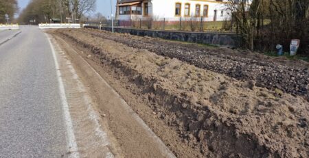 Am Straßenrand werden Straßenbauarbeiten durchgeführt, Erde und Kies werden verstreut, neben einem neu angelegten Weg. Im Hintergrund ist ein Haus mit Spitzdach und großen Fenstern zu sehen, umgeben von Bäumen. Der Himmel darüber ist weiterhin blau und klar.