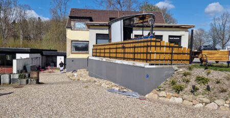 Ein zweistöckiges Haus mit braunem Dach und beiger Außenfassade wird gerade renoviert. Der Vorgarten mit Kies- und Steinbelag verleiht der Szene Farbe. Auf dem Haus ist eine Holzumzäunung angebracht, um die herum Baumaterialien und Schutt zu sehen sind. Im Hintergrund sind Bäume und ein klarer Himmel zu sehen.