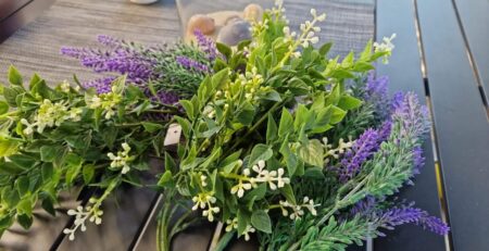 Eine Nahaufnahme eines Blumenarrangements mit lila Lavendel, weißem Schleierkraut und verschiedenen grünen Blättern. Der Strauß liegt auf einem Tisch mit einem grauen Tischset und einer dekorativen Vase mit neutral gefärbten Objekten im Hintergrund. Diese überraschende Blumengruß-Szene vermittelt ein rustikales, natürliches Gefühl.