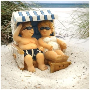 Zwei Kinderfiguren sitzen auf einem Strandkorb aus Korbgeflecht unter einem blau-weiß gestreiften Baldachin, eingebettet in Sand. In Badeanzügen und Sonnenbrillen wirken sie entspannt am Meer, umgeben von spärlichem grünem Gras. Im Hintergrund ist ein entfernter Meerblick zu sehen.Figur 'Urlauberpaar' im Strandkorb - weiss/blau