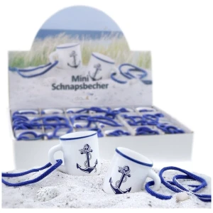 In einer Displaybox befinden sich mehrere Mini Schnapsbecher Stamperl 'Anker', weiß mit blauem Rand, jeweils verziert mit einem blauen Ankermotiv und blauen Schnüren. Der Hintergrund zeigt eine Strandszene mit Sand und Gras. Der Text auf der Box lautet „Mini Schnapsbecher“.