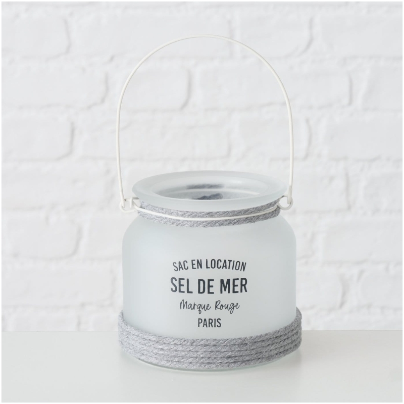 Exklusives Highline Windlicht Seldemer, Beige, Grau, Weiss Ein Milchglasgefäß mit einem grauen Seil um den Boden. Es hat einen weißen Griff und einen schwarzen Text mit der Aufschrift „Sac en Location Sel de Mer, Marque Rouge, Paris“. Der Highline Dekoaufsteller Saily steht vor einem weißen Backsteinmauerhintergrund.