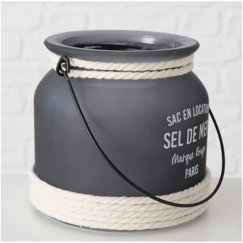 Exklusives Highline Windlicht Seldemer, Beige, Grau, Weiss Ein grauer Metallbehälter mit schwarzem Griff und weißem Seil, das oben und unten umwickelt ist. Er trägt den französischen Text „SAC EN LOCATION SEL DE MER Marque Rouge PARIS“. Dieser Highline Dekoaufsteller Saily steht vor einem weißen Ziegelhintergrund.