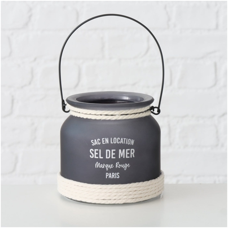 Exklusives Highline Windlicht Seldemer, Beige, Grau, Weiss Ein kleiner, runder, mattschwarzer Behälter mit Griff und weißem Seildetail an der Unterseite. Auf dem Behälter steht in Weiß der Text „SAC EN LOCATION SEL DE MER – Marque Rouge – PARIS“. Perfekt als Highline Dekoaufsteller Saily, hebt er sich elegant vom weißen Backsteinmauerhintergrund ab.