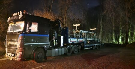 Ein großer schwarzer DAF-LKW parkt nachts auf einer unbefestigten Straße und transportiert einen Pritschenanhänger mit Metallkonstruktionen. Der mit Ausrüstung beladene Anhänger, der vom LKW und den umliegenden Lichtern beleuchtet wird, hat einfachen Zugang zu genügend Parkplätzen. Bäume und ein dunkler Himmel bilden den Hintergrund.