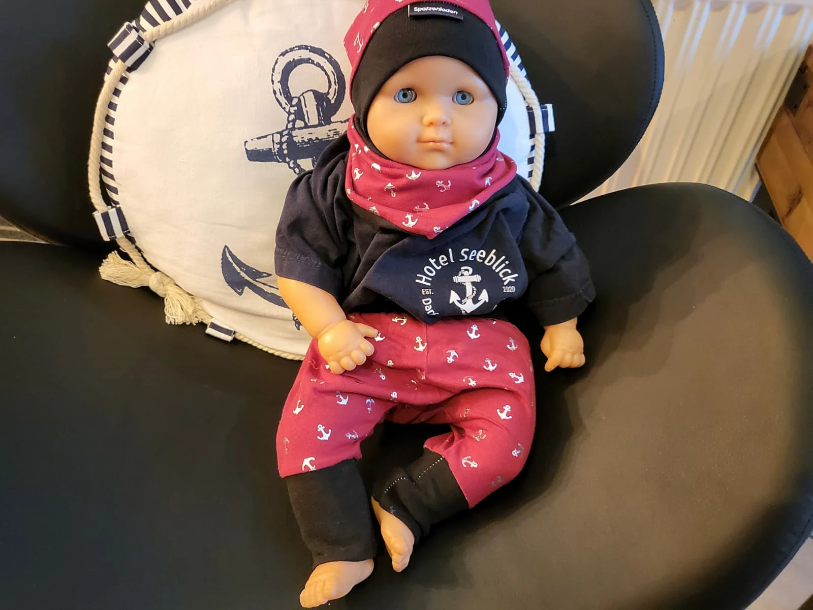 Eine Puppe, die mit Babyklamotten im maritimen Stil bekleidet ist, sitzt auf einem schwarzen Stuhl. Die Zuckersüß-Puppe trägt eine schwarze Beanie, ein marineblaues Hemd und eine rote Hose mit weißem Ankermuster. Hinter der Puppe befindet sich ein weißes rundes Kissen mit Ankermotiv und dem Text „Hotel Seeblick“.