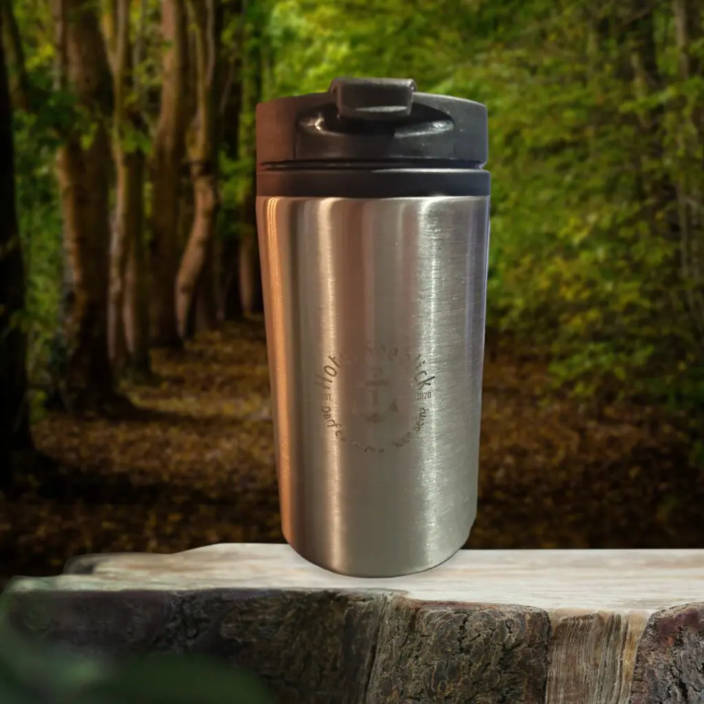 Ein Thermo-Reisebecher aus Edelstahl mit schwarzem Deckel steht auf einer Holzoberfläche. Der Hintergrund zeigt eine Waldszene mit hohen Bäumen und einem mit Laub bedeckten Weg. Der Becher sieht leicht abgenutzt aus, was auf häufigen Gebrauch hindeutet und die Essenz bewährter Thermobecher verkörpert.
