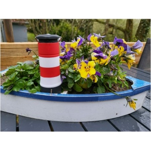 Ein kleiner blau-weißer Blumentopf in Bootsform steht auf einem schwarzen Gartentisch. Im Topf befinden sich leuchtend violette und gelbe Stiefmütterchen, zwischen denen sich ein kleiner rot-weiß gestreifter Leuchtturm befindet. In der Nähe verleiht eine LED-Taschenlampe mit 3 Funktionen der bezaubernden Szene einen modernen Touch, vor verschwommenem Grün und Holzelementen.