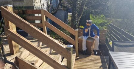 Auf einem Holzdeck befindet sich eine Treppe mit Handlauf. Am Fuß der Treppe sitzt auf einer Holzkiste die Statue eines Matrosen in blau-weißer Kleidung und hält sich ein Fernglas vors Auge. Die Tür zur Terrasse öffnet sich und gibt den Blick auf Bäume, einen Zaun und ein Haus im Hintergrund frei.