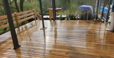 Eine nasse Holzterrasse mit einer Pergola aus Metall. Üppige, grüne Bäume und Pflanzen umgeben das Deck, und im Hintergrund ist ein überdachter Gegenstand zu sehen. Die Holzlatten des Decks glänzen vom Regen und sorgen für ein frisches und sauberes Aussehen.