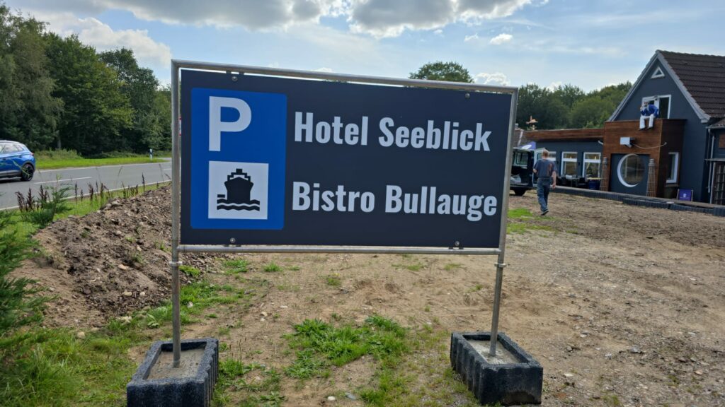 Auf einem Schild im Außenbereich sind die Informationen zum „Hotel Seeblick“ und zum „Bistro Bullauge“ mit einem Parkplatzsymbol (Hinweis: Parkplatz) zu sehen. Im Hintergrund sind eine Schotterfläche und ein Gebäudeteil zu sehen, außerdem ein blaues Auto und etwas Grün.