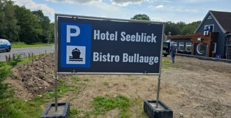 Auf einem Schild im Außenbereich sind die Informationen zum „Hotel Seeblick“ und zum „Bistro Bullauge“ mit einem Parkplatzsymbol (Hinweis: Parkplatz) zu sehen. Im Hintergrund sind eine Schotterfläche und ein Gebäudeteil zu sehen, außerdem ein blaues Auto und etwas Grün.