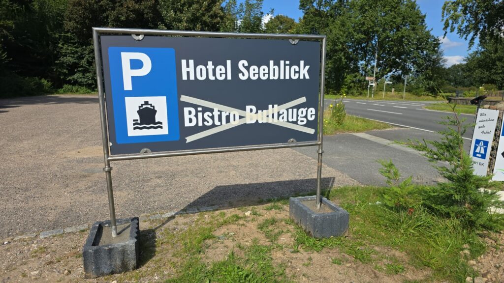 Am Straßenrand steht ein Schild in schwarz-blauem Design. Mit einem Park-P-Symbol, einem Schiffssymbol sowie den Aufschriften „Hotel Seeblick“ und „Bistro Bullauge“ dient es als hilfreicher Hinweis für Besucher. Das Schild wird von zwei Betonblöcken getragen, im Hintergrund sind Bäume und die Straße zu sehen.