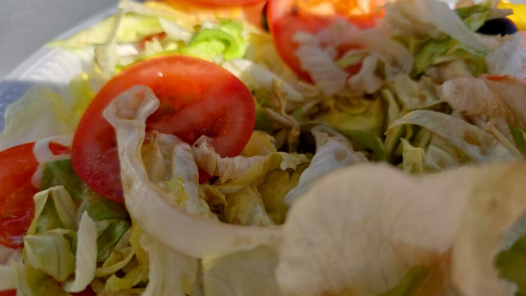 Nahaufnahme eines gemischten Salats mit geschnittenen Tomaten, Eisbergsalat und möglicherweise anderem Gemüse. Die Farben sind lebendig und heben die Frische der Zutaten hervor. Das Bild scheint bei natürlichem Licht aufgenommen worden zu sein, wodurch die Textur des Salats und der Tomaten hervorgehoben wird.