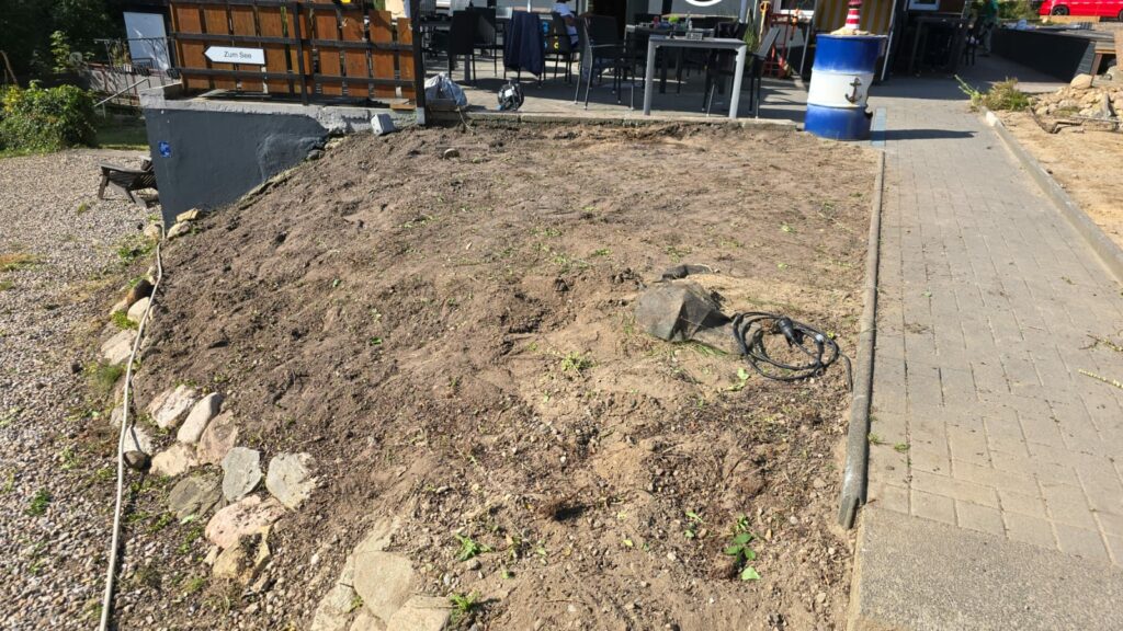 Ein kahler Fleck Erde auf einem abfallenden Hügel in der Nähe eines gepflasterten Gehwegs vor einem Gebäude stellt eine üble Fläche dar. Der Bereich ist durch eine Kombination aus Steinen und Beton begrenzt. Gartengeräte, darunter ein Schlauch, liegen verstreut auf dem Boden. Das Gebäude verfügt über Sitzgelegenheiten im Freien und Beschilderungen.