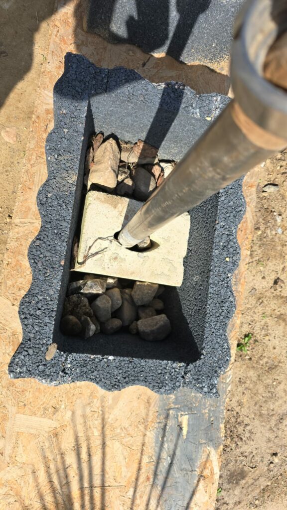 Ein rechteckiges Loch, das mit schwarzem Material ausgekleidet ist, enthält am Boden Steine. Ein Metallpfosten ist in einem Betonblock im Loch verankert. Der umgebende Boden, der an Der Busch, der nicht sein darf erinnert, scheint eine Mischung aus Erde und Sperrholz zu sein.