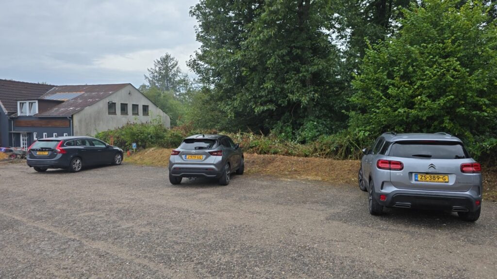 Drei parkende Autos auf einem unbefestigten Parkplatz, umgeben von Bäumen und Sträuchern, mit einem Gebäude im Hintergrund.