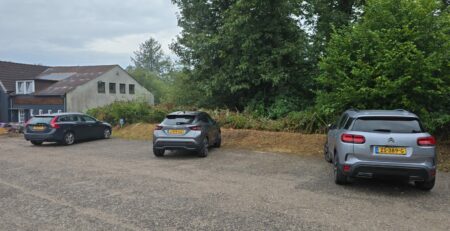 Drei parkende Autos auf einem unbefestigten Parkplatz, umgeben von Bäumen und Sträuchern, mit einem Gebäude im Hintergrund.