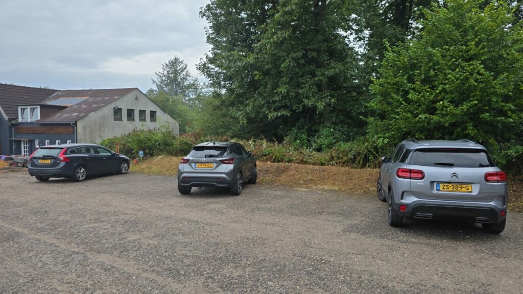 Drei parkende Autos auf einem unbefestigten Parkplatz, umgeben von Bäumen und Sträuchern, mit einem Gebäude im Hintergrund.