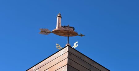 Wetterfahne aus Kupfer mit einem Haus und Pfeil auf einem Holzgebäude, vor blauem Himmel. Dekorative Architekturdetails.