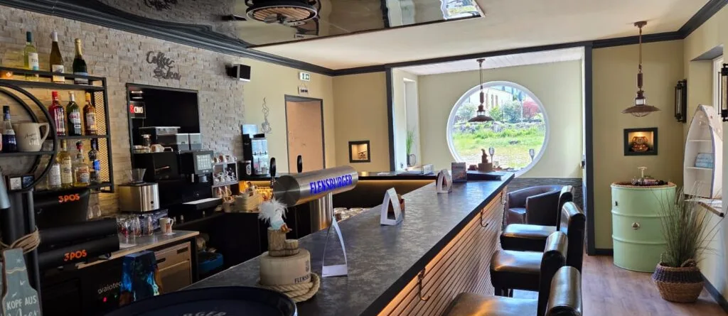 Inneneinrichtung einer Bar mit einer langen Theke und schwarzen Stühlen in einem hellen Raum.