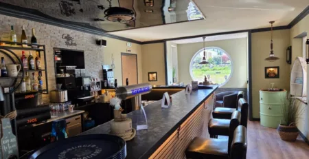 Inneneinrichtung einer Bar mit einer langen Theke und schwarzen Stühlen in einem hellen Raum.