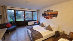 Ein gemütliches Schlafzimmer in Flensburg mit einem großen Bett mit weißer und grauer Bettwäsche. Eine graue Couch mit roten Kissen steht neben einem großen Fenster. Über dem Bett hängt ein fünfteiliges Kunstwerk mit einem Sonnenuntergang über dem Meer. Die hellen Holzböden und der Blick ins Grüne machen es zu einem echten Geheimtipp.