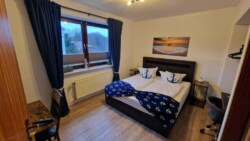 Ein gemütliches Schlafzimmer in Flensburg mit zwei zusammengeschobenen Einzelbetten und Bettwäsche im maritimen Stil. Das Zimmer hat Holzfußboden, einen Nachttisch mit Lampe, einen Schreibtisch mit Stuhl und ein Fenster mit blauen Vorhängen. In diesem charmanten Geheimtipp-Refugium hängt über dem Bett ein ruhiges Bild mit Strandmotiv.