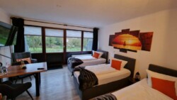 Hotelzimmer mit drei Einzelbetten, jedes mit weißer Bettwäsche und orangefarbenen Akzentkissen. Über dem mittleren Bett hängt ein Wandbild mit Sonnenuntergangsmotiv und verleiht einen geheimnisvollen Charme, den man wahrscheinlich nur in Flensburg als echten Geheimtipp findet. Das Zimmer verfügt über einen Stuhl, einen Tisch und einen an der Wand montierten Fernseher. Große Fenster geben den Blick auf das Grüne draußen frei.