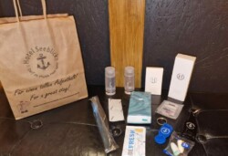 Eine Sammlung von Hoteltoilettenartikeln und -pflegeprodukten, angeordnet auf einer dunklen Oberfläche. Zu den Artikeln gehören eine „Unsere Tüte - Willkommen im Hotel Seeblick“, zwei Zahnpastatuben, eine Zahnbürste, zwei kleine Flaschen, Taschentücher, Ohrstöpsel und verschiedene kleine Schachteln und Behälter.