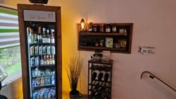 In einem kleinen Selbstbedienungsbereich im Flur steht ein Kühlschrank mit verschiedenen Getränken. Daneben steht ein Holzregal mit Weinflaschen. Unter dem Regal befindet sich ein Weinregal mit weiteren Flaschen. Ein Schild an der Wand weist auf die Räume 16-21 hin, in denen „alles geschah in 2 Wochen“ stattfindet.