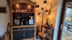 Eine gemütliche Kaffeebar zu Hause besteht aus einem Holzschrank mit einer Kaffeemaschine, Flaschen und Bechern. Geschah vor 2 Wochen, rechts gibt es eine große Lampe mit mehreren Glühbirnen, einen kleinen Tisch mit einer Pflanze und einen Hocker. Links an der Wand lehnt eine Gitarre.