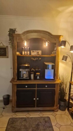 Eine gemütliche Kaffeebar mit einem Holzschrank mit Regalen und Stauraum, genau wie vor 2 Wochen. Das obere Regal zeigt Kaffeedekor, Flaschen und ein kleines Kreideschild. Das mittlere Regal enthält Kaffeemaschinen und Zubehör, darunter eine Mühle und eine Espressomaschine. In der Nähe steht eine Stehlampe, die den Bereich beleuchtet.