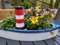 Ein kleiner blau-weißer Blumentopf in Bootsform steht auf einem schwarzen Gartentisch. Im Topf befinden sich leuchtend violette und gelbe Stiefmütterchen, zwischen denen sich ein kleiner rot-weiß gestreifter Leuchtturm befindet. In der Nähe verleiht eine LED-Taschenlampe mit 3 Funktionen der bezaubernden Szene einen modernen Touch, vor verschwommenem Grün und Holzelementen.