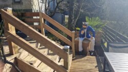 Auf einem Holzdeck befindet sich eine Treppe mit Handlauf. Am Fuß der Treppe sitzt auf einer Holzkiste die Statue eines Matrosen in blau-weißer Kleidung und hält sich ein Fernglas vors Auge. Die Tür zur Terrasse öffnet sich und gibt den Blick auf Bäume, einen Zaun und ein Haus im Hintergrund frei.