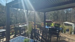 Eine sonnenbeschienene Außenterrasse mit mehreren schwarzen Tischen und Stühlen unter einer Pergola. Im Hintergrund sind Bäume und Grünflächen zu sehen. Rechts befindet sich eine Holztür, die zu einem Gartenbereich führt, über dem auf einem Schild „HOTEL SERLICK“ steht.