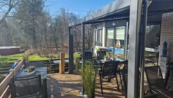 Eine geräumige Außenterrasse mit Holzbelag verfügt über mehrere schwarze Tische und Stühle, umgeben von grünen Pflanzen und einigen Topfblumen. Die Terrasse ist teilweise von einer Pergola überdacht. Im Hintergrund befindet sich ein üppiger, grasbewachsener Hof, der von Bäumen und einem roten Schuppen gesäumt ist.