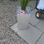 Ein hoher, rechteckiger Pflanzkübel mit Grünpflanzen steht auf einer Kies- und Pflasterfläche. Am Pflanzkübel ist ein kleines rundes Schild angebracht, das einen roten Kreis mit einer diagonalen Linie zeigt, die über einem Vogel verläuft. Es weist darauf hin, dass das Füttern von Vögeln verboten ist. Bitte lassen Sie Haustiere nicht in der Nähe der Pflanzen pinkeln. Ein hoher, rechteckiger Pflanzkübel mit Grünpflanzen steht auf einer Kies- und Pflasterfläche. Am Pflanzkübel ist ein kleines rundes Schild angebracht, das einen roten Kreis mit einer diagonalen Linie zeigt, die über einem Vogel verläuft. Es weist darauf hin, dass das Füttern von Vögeln verboten ist. Bitte lassen Sie Haustiere nicht in der Nähe der Pflanzen pinkeln.