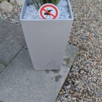 Ein hoher, grauer Blumentopf mit weißen Kieselsteinen und einer grünen Pflanze steht auf einer Kiesfläche. Ein Schild auf dem Blumentopf zeigt eine schwarze Hundesilhouette mit einem roten Verbotskreis darum, der darauf hinweist, dass Hunde in diesem Bereich nicht pinkeln dürfen. Ein hoher, grauer Blumentopf mit weißen Kieselsteinen und einer grünen Pflanze steht auf einer Kiesfläche. Ein Schild auf dem Blumentopf zeigt eine schwarze Hundesilhouette mit einem roten Verbotskreis darum, der darauf hinweist, dass Hunde in diesem Bereich nicht pinkeln dürfen.